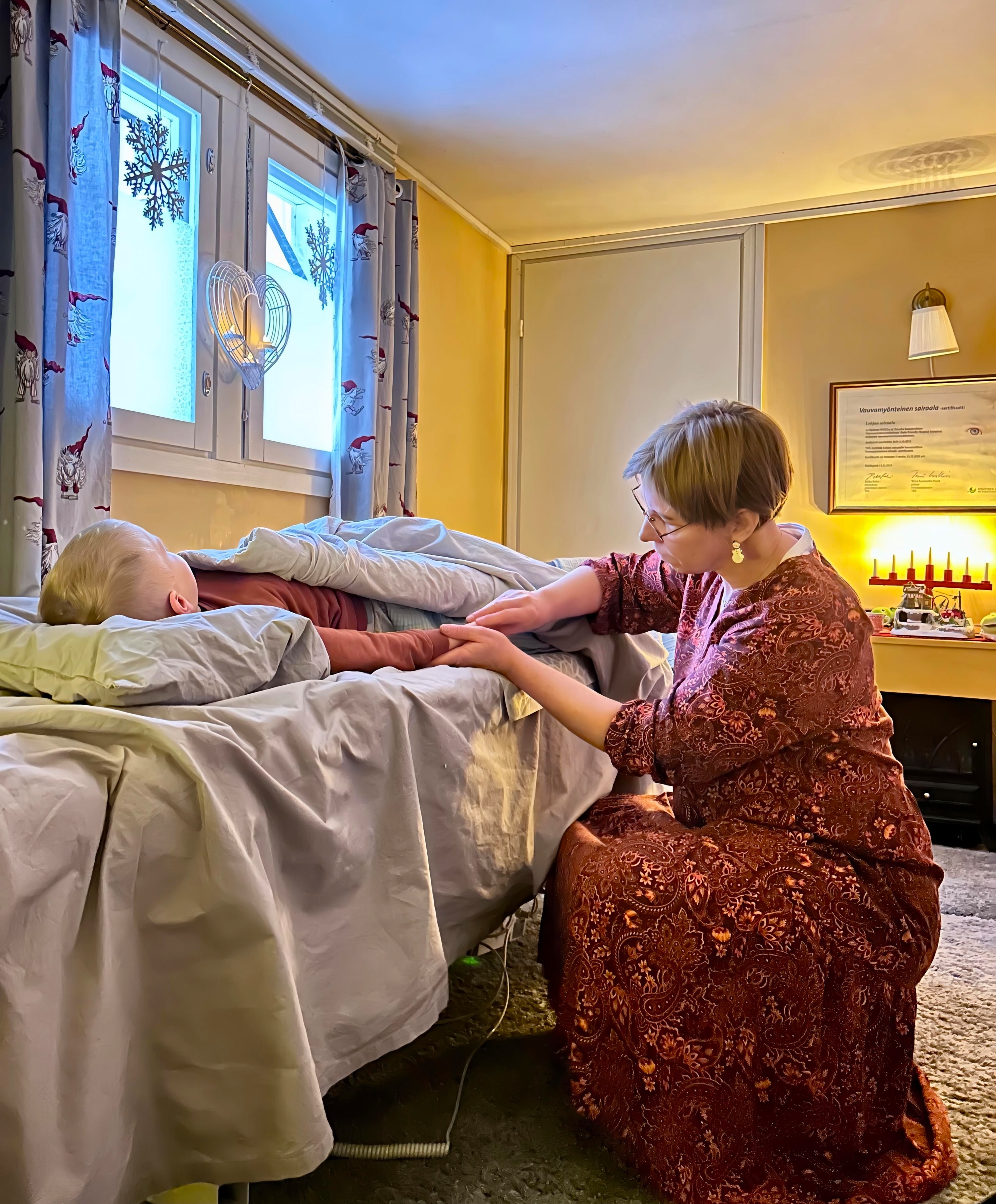 Vyöhyke terapeuttinen lapsen hieronta. Hoitaja mekko päällä kyykyssä hieroo lapsen kättä. Lapsi harmaan peiton alla rauhallisena. Tunnelmallinen huone, tonttu verhot. 