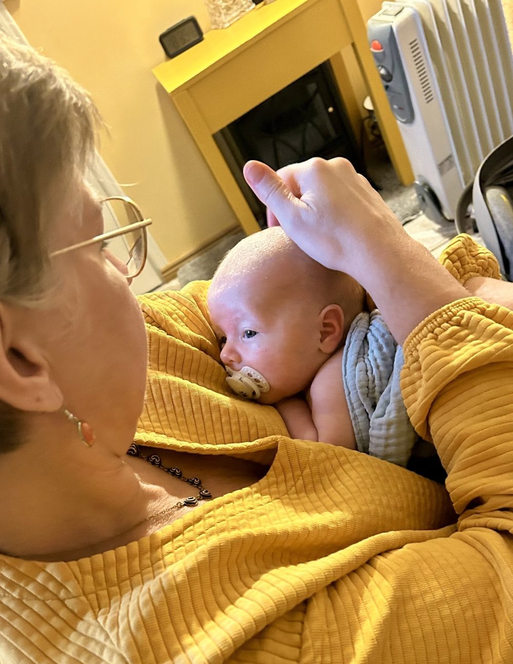 Titta hieroo vauvaa keltainen paita päällään tuolilla istuen. vauva sylissä ilman vaatteita, kietoutuneena siniseen pyyhkeeseen. Titta hieroo kevyesti vauvan päätä samalla vauvaa katsoen.