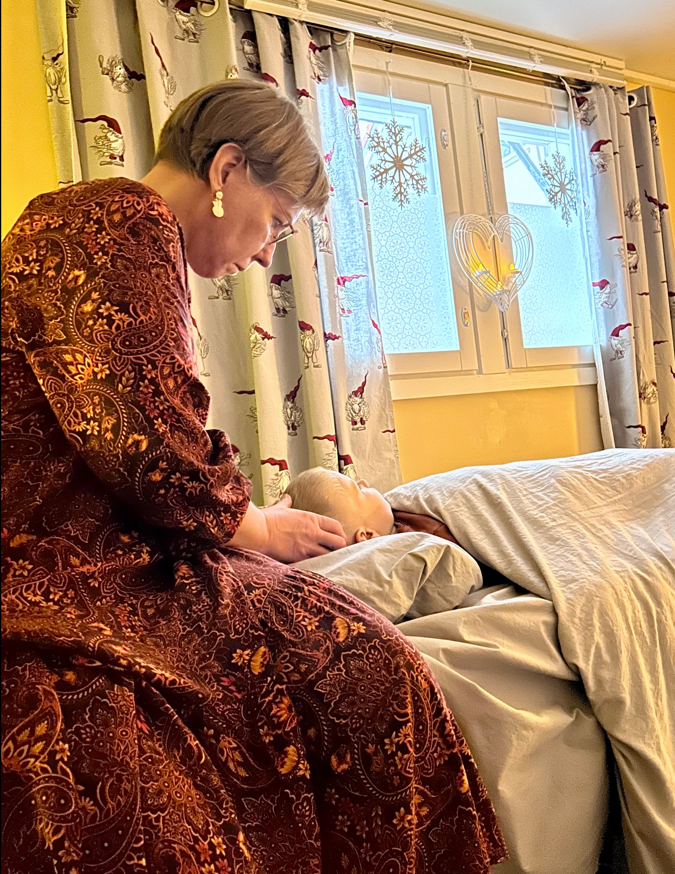 Vyöhyketerapia lapselle. Vyöhyketerapeutti on mekko päällä ja hieroo lempeästi lapsen päätä lapsen maassa hoito pöydällä harmaan peiton alla. Tunnelma on lempeä. 