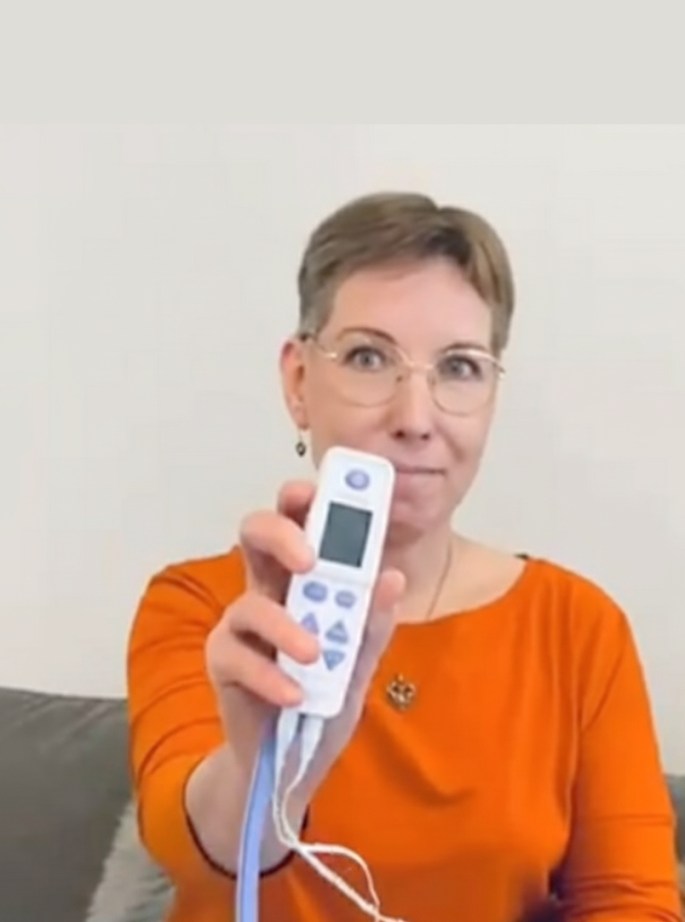 Nainen pitää kädessään TENS+ -kivunlievityslaitetta, pukeutuneena oranssiin puseroon ja katsoo suoraan kameraan.