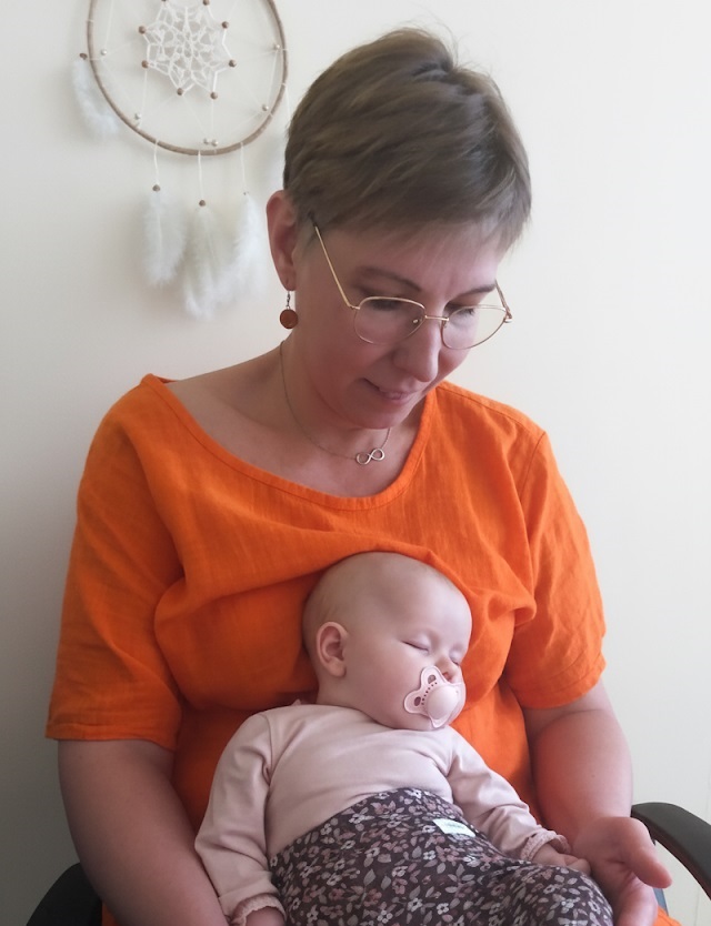 vauva Titta järvenpään sylissä. Titalla oranssi paita ja vauvalla vaaleanpunaiset vaatteet. Vauvalla tutti suussa ja hän nukkuu