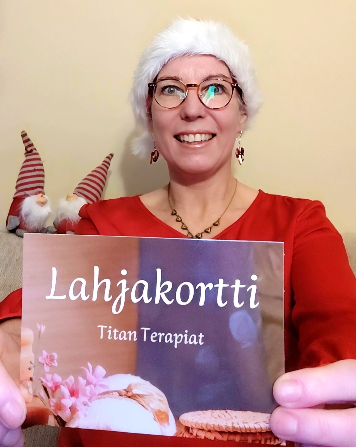 Henkilö punaisessa paidassa ja joulupipossa , tonttulakissa, pitää kädessään lahjakorttia, jossa lukee 'Lahjakortti Titan Terapiat'. Taustalla näkyy jouluisia koristeita.