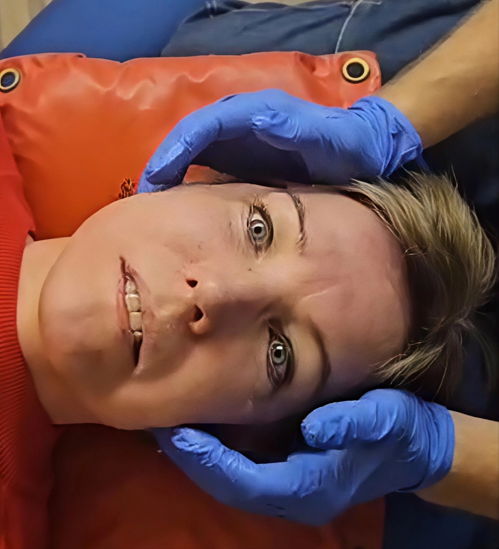 Kuvassa näkyy henkilö makamassa oranssilla tyynyllä, kun toinen henkilö käyttää sinisiä käsineitä hänen kasvonsa hierontaan tehden purentalihashierontaa