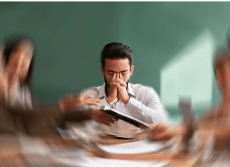 Mies istuu neuvottelupöydän ääressä, pitelee kasvojaan ja näyttää mietteliäällä ilmeellä.